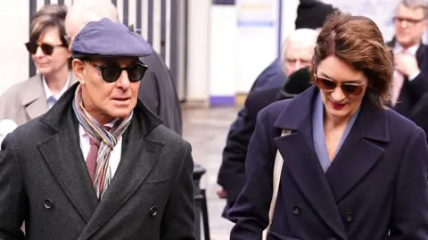 PA Media Stanley Tucci and Felicity Blunt arriving at the memorial. Both wear sunglasses and formal clothing