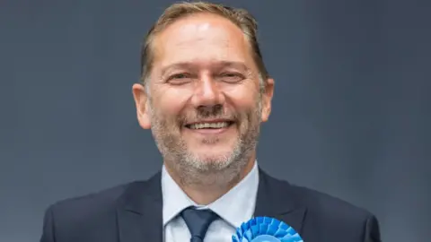 A man with short dark hair smiles. He is wearing a business suit and a blue rosette. 
