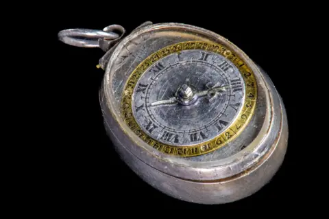 The Cromwell Museum A silver-coloured oval-shaped 17th Century watch. The clock face is round and set into the top, with a gold rim etched with numbers and a silver interior with arms. It has a hanging loop.
