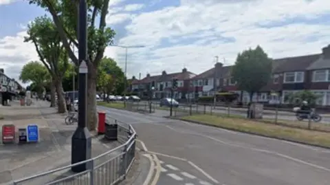 Google A dual carriageway, flanked by a parade of shops on one side and terraced housing on the other. 