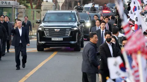 Reuters South Korean impeached President Yoon Suk Yeol walks outside the Seoul detention center after his release, in Uiwang, with crowds of supporters waving South Korea flags and a police convoy behind him.