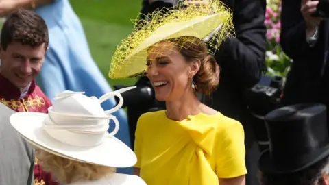 PA Media Catherine, Princess of Wales, is pictured among crowds at Royal Ascot wearing a yellow dress and hat. She is laughing. 