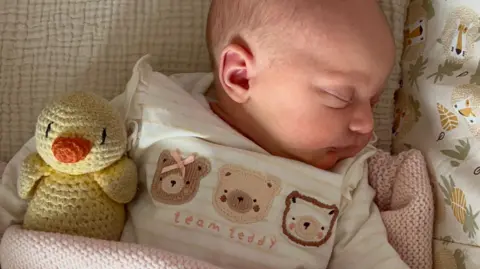 Maddie Biggs A baby girl sleeps in a cot. She is wearing a sleepsuit in pink tones that says 'Team Teddy' with a teddy bear motif and has a knitted duck under her arm. 