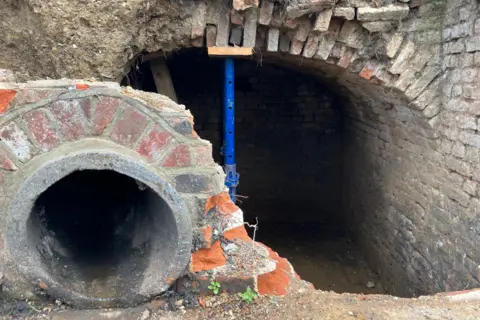 Henry Godfrey-Evans/BBC A view from outside of the tunnel with the exit of a sewage pipe to the side. There's an arch made of loose bricks and a metal post propping up some bricks.
