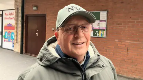 Vikki Irwin/BBC Brain Wisker is wearing a grey green coat and hat. The hat has the letters "NY" on them. He is wearing glasses and smiling at the camera.