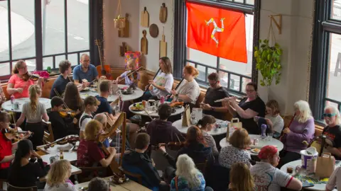 A group of people sat around tables playing harps and flutes and guitars. A manx Flag flies in the window. 