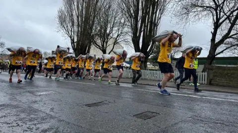 BBC A group of people running on a road carrying bags on coal on their backs. They are wearing yellow t-shirts and black shorts. 