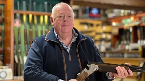 Andy Whitemore is wearing a blue and white check shirt, a blue zip-neck jumper and a navy blue fleece gilet with brown leather piping and is holding a shotgun in his shop with a blurred background of guns