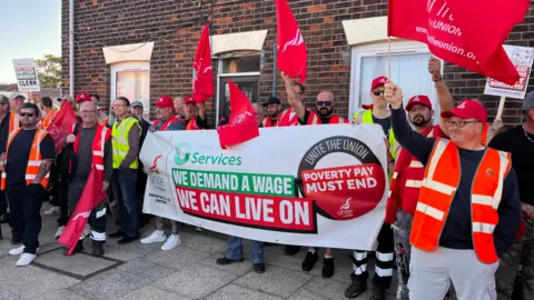 Martin Giles/BBC A row of workers on strike as part of a picket line. Some are waving flags and others are holding a banner which say, WE DEMAND A WAGE WE CAN LIVE ON. POVERTY PAY MUST END.