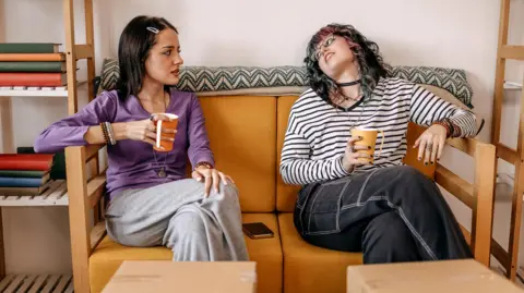 Two women holding mugs sit on a sofa with cardboard boxes in front of them, and shelves either side of them, with some books on the shelves.