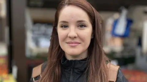 Martin Giles/BBC Maria Agachi smiles at the camera as she is photographed at a market. She is wearing a black rain jacket and brown backpack. She has long brown hair. 