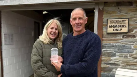Becca and Steve Peck are smiling while  standing in front of a stone building. Becca woman is wearing a green quilted jacket, and Steve is dressed in a navy blue knitted sweater. They are both holding a clear glass trophy.