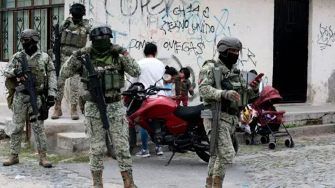 Getty Images Four Mexican army soldiers stand guard. They are armed with assault rifles, as well as wearing full military gear and face mask.