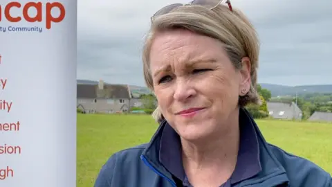 Debbie Dixon has short blonde hair and has glasses resting on her head. She is wearing a dark blue jacket and standing with a field behind her. On the left is part of a white sign for Manx Mencap.