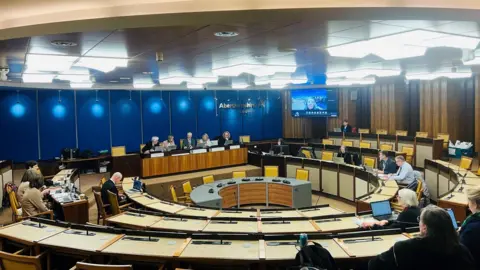 BBC A meeting of the Integration Joint Board (IJB) - the body which runs health and social care services in the region - with people sitting in a meeting room, under bright lights, with an Aberdeenshire Council sign on wall.