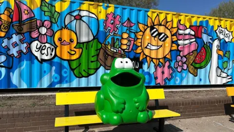 Nottingham City Council A green frog-shaped litter bin sat on a bench, with a summer-themed mural on a wall behind it.