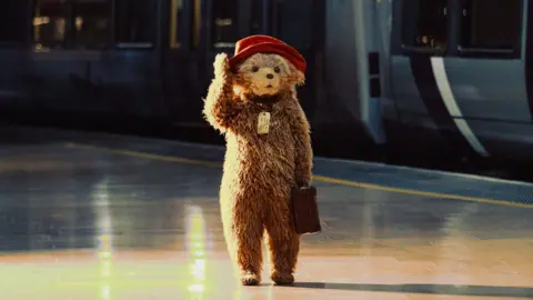 PA Small bear with red hat and suitcase stands alone on a train platform beside a parked train