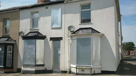 An image of a terraced street in Blackpool.  There is a Rydal Avenue sign on the road.  Two of the houses are boarded up with metal plates on their windows and doors.  It is a sunny day