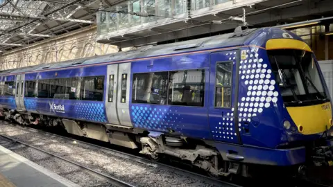 PA Media A blue liveried ScotRail trainsits at a platform. We can see three carriages.