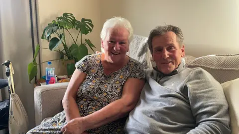 The image shows a woman and a man sitting on their sofa, smiling at the camera.