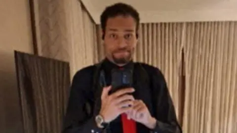 A man wearing a black button-down shirt and red tie holds a phone as he takes a photo of himself