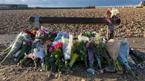 Lucinda Adam/BBC A pile of flowers left by a wooden post next to a shingle beach.