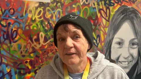 A woman wearing a black hat and grey hoodie standing in front of a multi-coloured painting on a wall