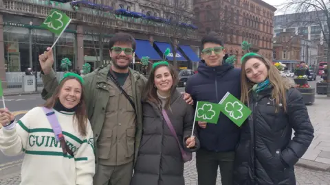 Sekelompok lima orang muda - tiga wanita dan dua pria - berpose untuk foto di Belfast tengah. Tiga dari mereka memegang bendera hijau kecil dengan simbol shamrock. Para pria mengenakan kacamata berbingkai hijau dan para wanita mengenakan ikat rambut hijau berbulu dengan simbol shamrock yang menonjol dari pita. 