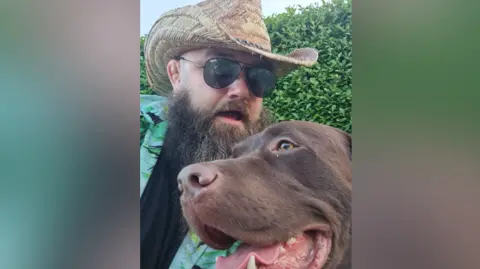 Philip and his dog - a chocolate Labrador. Philip wears a straw hat, aviator sunglasses and a Hawaiian shirt. 