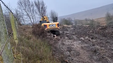 Roger Dobson A yellow JCB work vehicle is working on a muddy path that is bordered by shrubs on the right and a fence made of wooden fence posts and metal wires on the left. The front of the work vehicle is obscured by a tall brown shrub but it appear to have a digger on its front and continuous track wheels. There are fells in the distance.