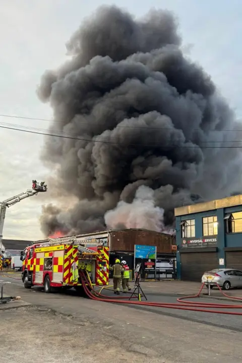 Bedfordshire Fire and Rescue Service A huge tower of dark black smoek rises from a building, dominating the top two thirds of the image, while a fire engine and crews are working in the bottom third.