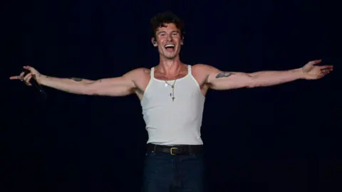 Getty Images Shawn Mendes stands on stage smiling with his arms open wearing a white vest top and jeans.