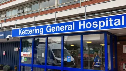 Sam Read/BBC The blue framed entrance to Kettering General Hospital with a blue sign above the door. A security guard stands inside the entrance. 