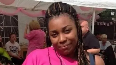 Facebook Lianne Gordon is smiling from the shoulders up. She is wearing a pink top and people sitting under the tent can be seen behind her