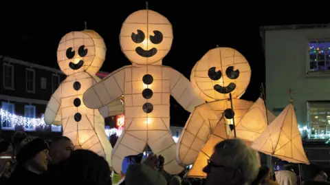 Three giant gingerbread men lanterns have been lit up. There are Christmas lights in the background. 