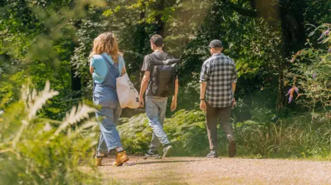 Three people walking on a gravel footpath through the Forest of Dean. On either side of the path there are trees, buses, and flowers.