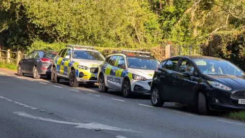 Eddie Mitchell Two police cars and two black vehicles parked beside a road with trees in the background. The photo is taken from the opposite side of the road