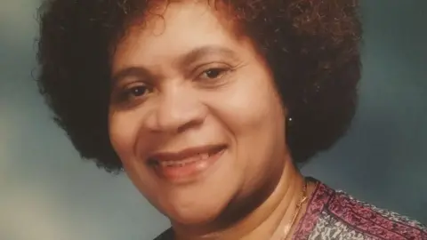 Leeds City Council A woman with short dark afro hair, wearing a pink and white patterned top.