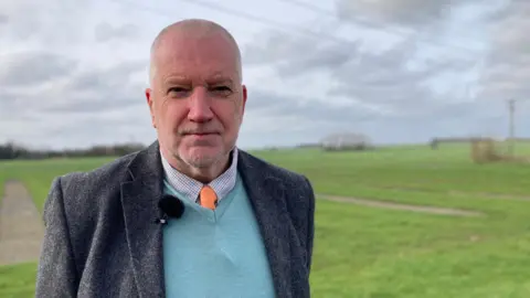 A man in a grey blazer, a blue jumper, a shirt and an orange tie is smiling into the camera with a microphone on his shirt. The background is blurred but a green field can be seen under a grey cloudy sky.