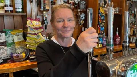The photo shows a woman standing behind a bar in a pub. She has brown hair, and is wearing a black jumper. She's holding a pump and is about to pull a pint. She's standing in front of bottles, packets of crisps and glasses. 