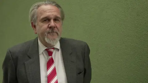 Beckinsale-Yates is making a neutral face and looking towards the camera. He has medium length grey hair. He is stood behind a green wall and is wearing a suit and tie. 