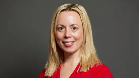 UK Parliament Parliamentary portrait of Sarah Hall, the Labour MP for Warrington South. She has mid-length blonde hair and a red blouse on.