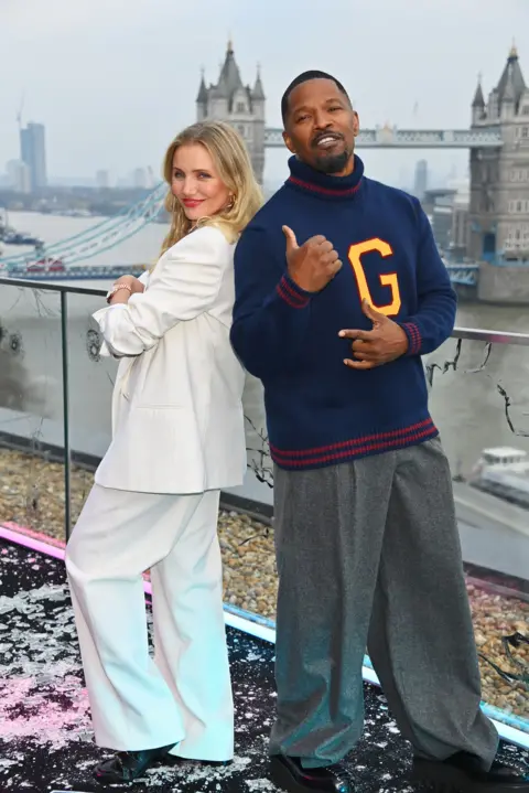 Getty Images Cameron Diaz, wearing a bright white suit, and Jamie Foxx, wearing a blie jumper a grey smart trousers, smile together on a balcony overlooking London's Tower Bridge as part of a publicity event for their new film. The film's logo is on the floor.