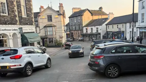 A black car being driven the wrong way up the main street in Ambleside, Lake District. There are cars driving in the oncoming direction. There are lots of shops and buildings which include pubs, outdoor shops and a gallery. 