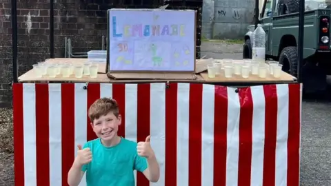 A picture of Hugo smiling with his thumbs up. He is stood in front of a red and white striped lemonade stand. Cups of lemonade are on the table, with a sign that reads 'LEMONADE 30P'