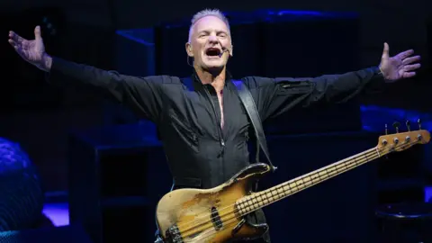 The image captures Sting centre stage, guitar strapped across his front, arms stretched wide in an iconic pose. He stands beneath the lights and head microphone in place.