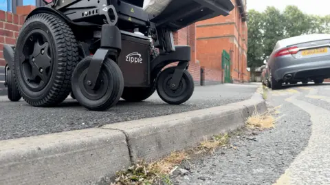 A close up of the wheels of a motorised wheelchair, sitting on the edge of a pathway next to double yellow lines