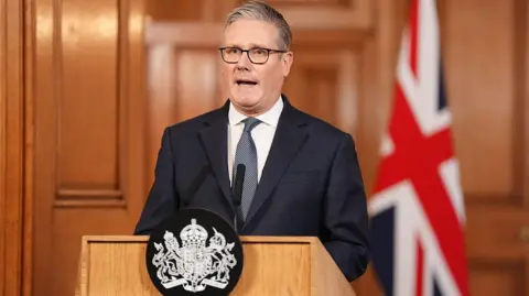 Starmer pictured standing in front of a lecturn in Downing Street while giving a speech. He is wearing a dark suit and tie and is standing in front of a flag of the United Kingdom which can be seen in the background. 