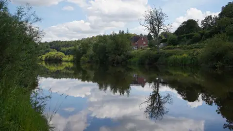 The River Severn has trees and bushes on each side. To the left there are green fields with some red brick properties seen in the distance. 