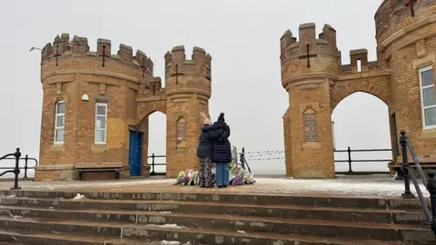 Two people stand in front of flower tributes left near the beach close to where they went into the water